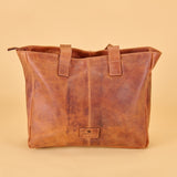A distressed brown leather tote bag with dual top handles and a small rectangular brand plaque on the front, set against a solid beige background.
