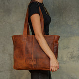 A woman in a black t-shirt carries a large brown distressed leather tote bag with long shoulder straps, shown from the side against a textured gray background.