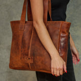 A person holding a large brown leather tote bag with distressed texture. The bag has two shoulder straps, a vertical seam, and a small rectangular logo patch near the bottom front.