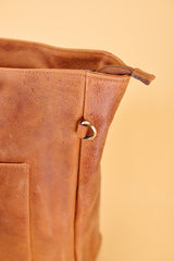 Close-up of a brown leather bag corner showing a zipper pull, metal ring detail, and a side pocket against a warm beige background.