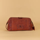 A brown leather toiletry bag with a front patch logo and side strap, photographed against a solid beige background.