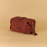 A brown leather toiletry bag with a top zipper and a small rectangular logo patch, photographed on a solid beige background.