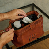 A person's hands are shown placing toiletry bottles into a brown leather travel pouch bag resting on a glass-topped table.