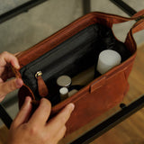 A brown leather travel pouch or toiletry bag with a black interior, shown being held open by two hands over a glass table.