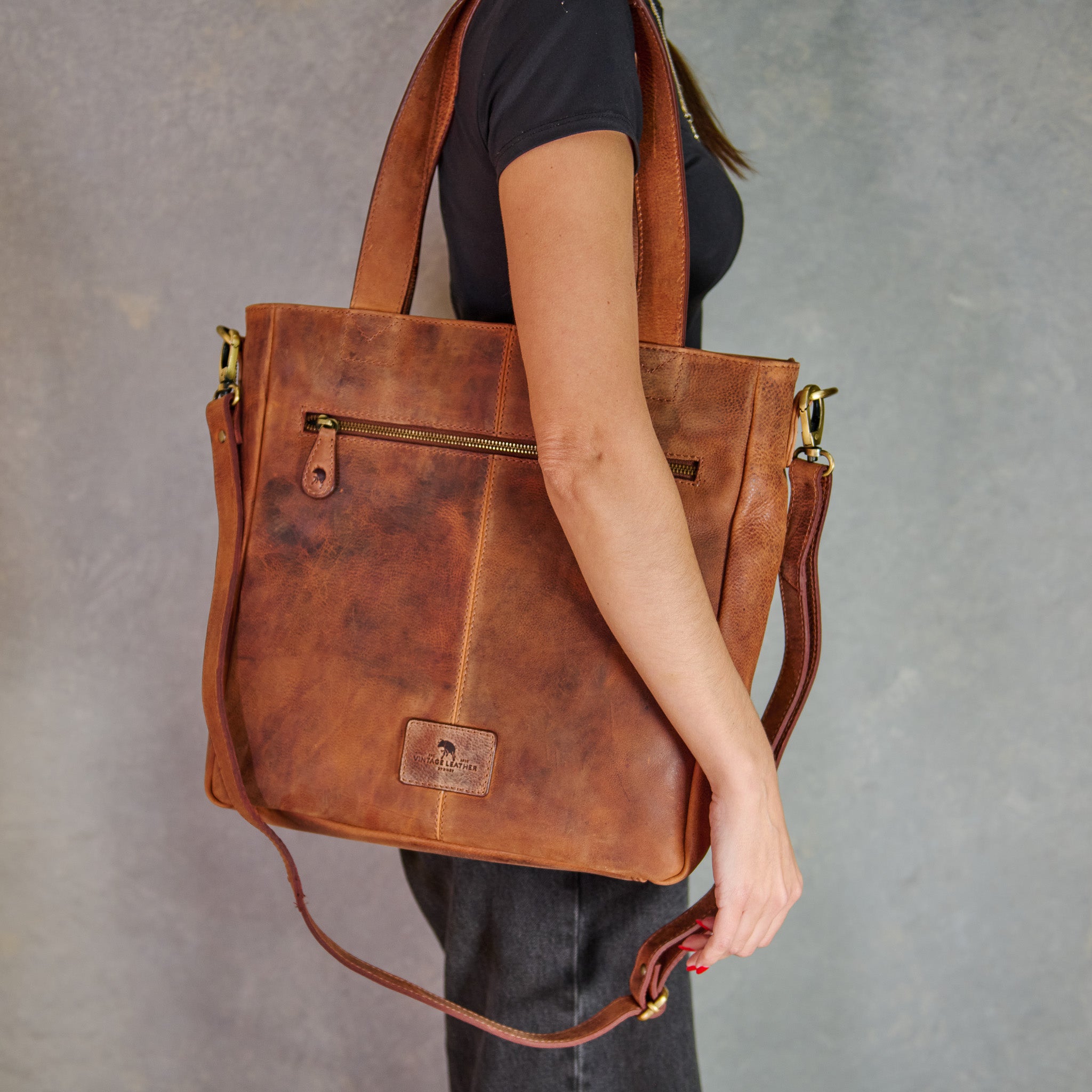 A person wearing a black t-shirt and dark jeans carries a large brown distressed leather tote bag with gold-tone hardware against a textured gray studio background.