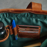 Close-up of the interior of a brown leather bag with a dark green satin lining. A brown leather card holder pocket contains several cards, a blue pen is tucked into a side loop, and part of a black bottle is visible on the left.