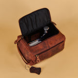A brown distressed leather travel or toiletry bag with its lid open, showing a black interior and a perfume bottle inside, set against a solid beige background.