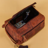 A brown distressed leather toiletry bag with an open top zipper, shown with its lid propped open and a small leather tag hanging from the zipper pull. The bag is filled with toiletry items and photographed on a solid beige background.