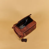 A brown leather toiletry bag with an open top zipper, shown on a solid beige background. Inside the bag, a small dark bottle is visible.