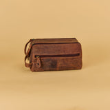 A brown distressed leather zippered toiletry bag with a top handle, shown on a solid beige background.