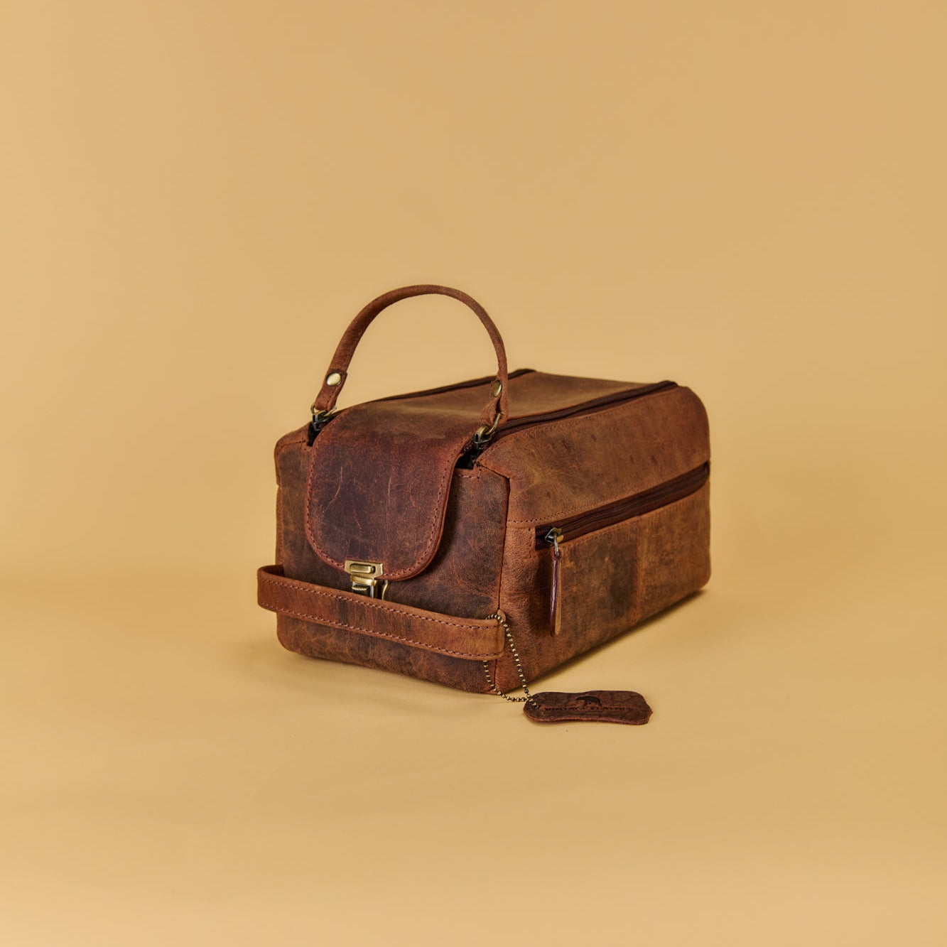 A dark brown distressed leather travel case with a top handle, front flap pocket, and zippered side pocket, shown on a solid beige background.