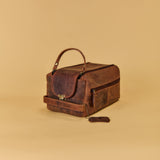 A dark brown distressed leather travel case with a top handle, front flap pocket, and zippered side pocket, shown on a solid beige background.