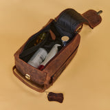 A brown distressed leather toiletry bag shown open, revealing a black interior lined with protective padding and containing travel-sized toiletry bottles. The bag is photographed on a solid warm beige background.