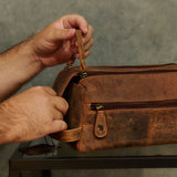 A person's hands holding and demonstrating the top handle of a distressed brown leather toiletry bag resting on a glass table against a textured gray wall.