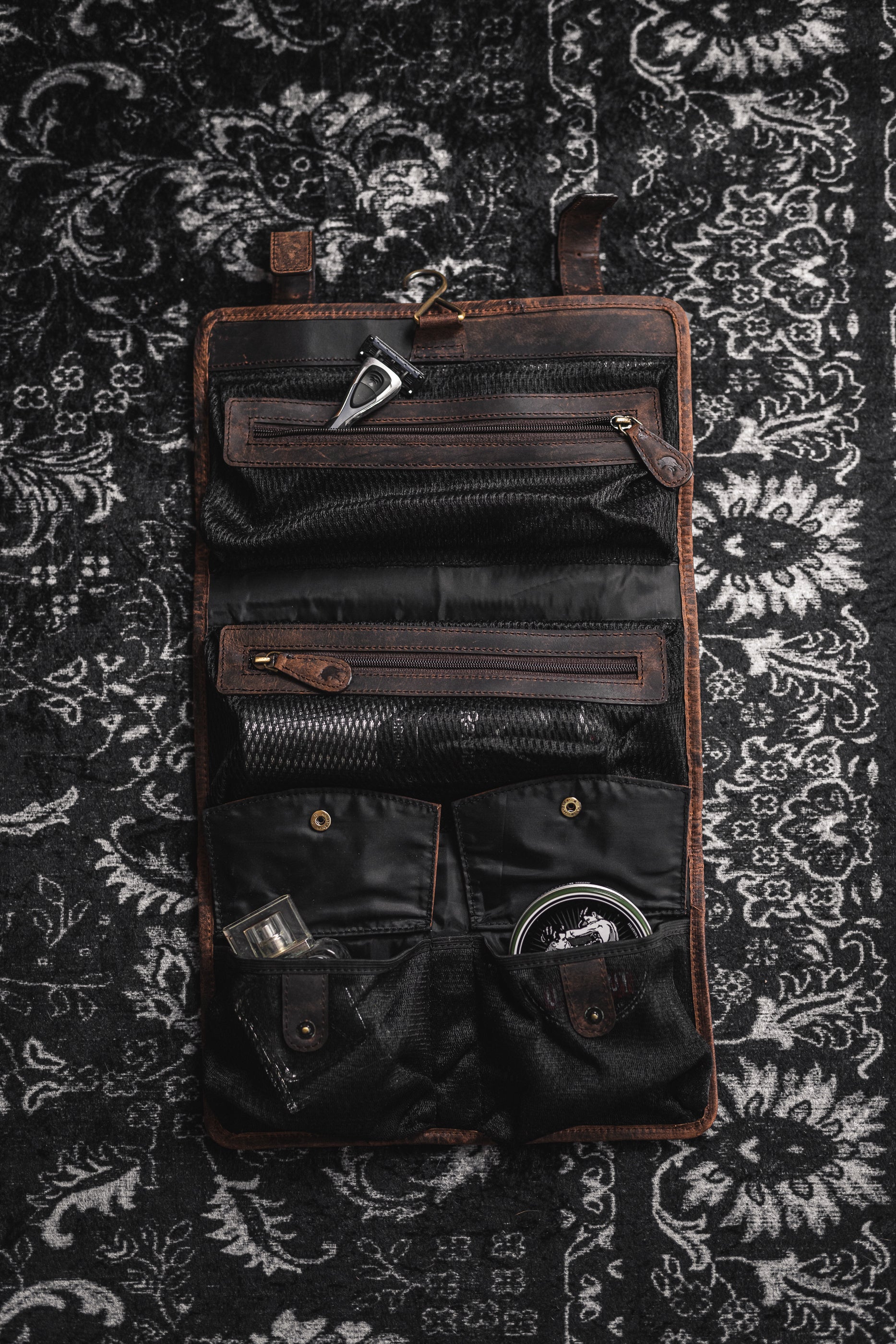 The Leather Hanging Toiletry Bag Rio by Vintage Leather Sydney, in brown and black, sits open on a patterned black and white rug, showcasing compartments filled with grooming items—an Italian-inspired example of elegance.