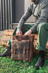 A person in a gray turtleneck, green pants, and black boots sits on a brick ledge holding the Vintage Leather Laptop Backpack Marcus with reinforced stitching, stylish zippers, and buckles on the grass.
